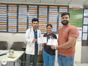 Two people receiving their certificates from a doctor in front of the departmental notice board