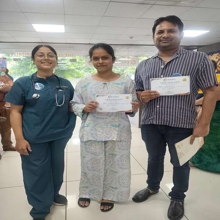 Two adults holding their certificates of participation, standing with a doctor near the hospital entrance